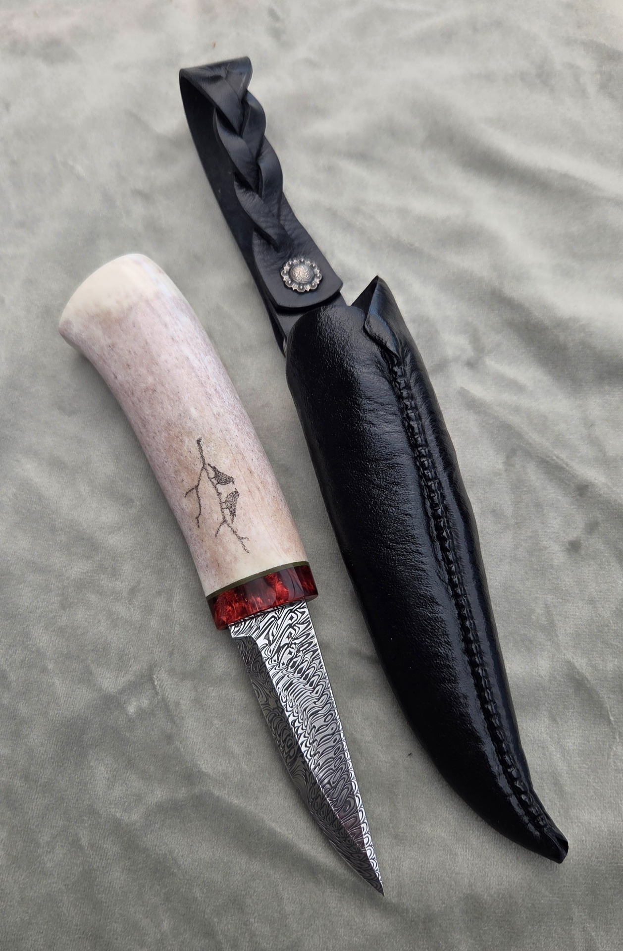 2026 "Glaskogen 12/2026" Rostfritt damaskus blad av Ingemar Nordell. Skaft av älghorn med front av stabiliserad masurbjörk, detalj av vulkanfiber. Mönster av scrimshaw (nålspetsgravyr).